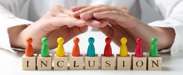 Image of Woman protecting colorful pawns and wooden cubes with word Inclusion at white table, closeup. Banner design