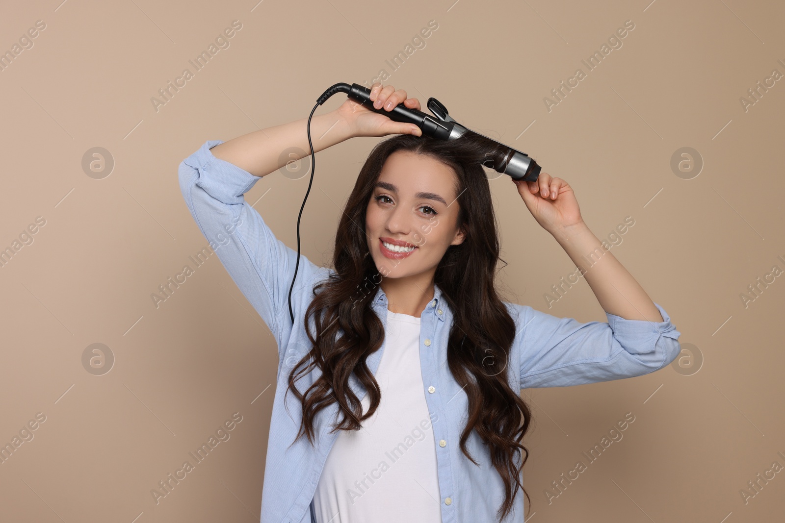 Photo of Happy woman using curling hair iron on beige background