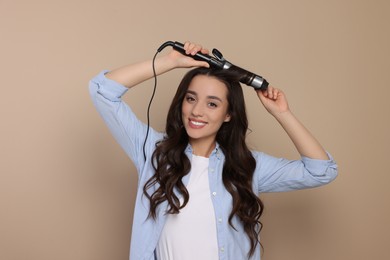 Happy woman using curling hair iron on beige background