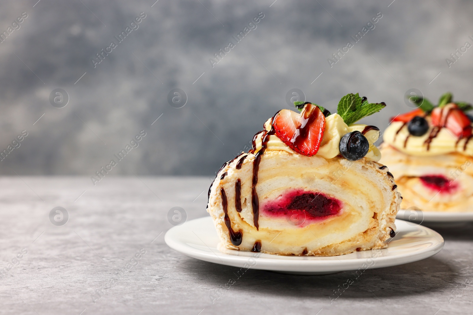 Photo of Pieces of tasty meringue roll with jam, cream, strawberry, blueberry and mint on light grey table, closeup. Space for text