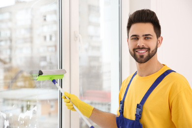 Photo of Professional young janitor in uniform cleaning window indoors