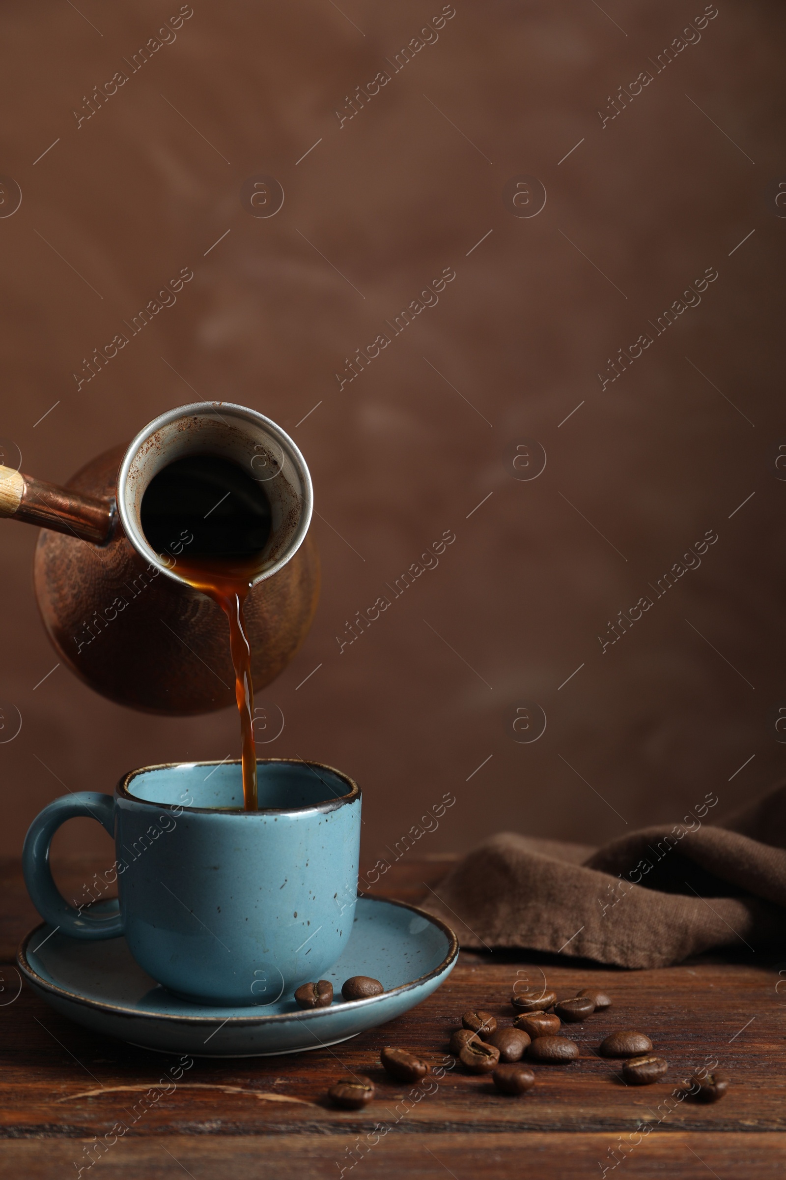 Photo of Turkish coffee. Pouring brewed beverage from cezve into cup at wooden table against brown background, space for text