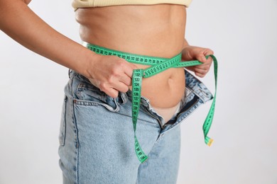 Woman in unfit jeans measuring her waist on light background, closeup. Weight loss concept