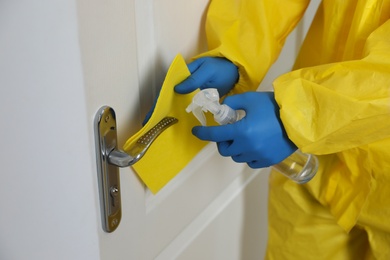 Photo of Employee in protective suit and gloves sanitizing door knob indoors, closeup. Medical disinfection