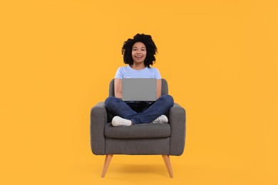 Happy young woman with laptop sitting in armchair against yellow background