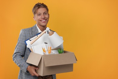 Photo of Happy unemployed young man with box of personal office belongings on orange background. Space for text