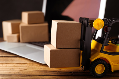 Photo of Toy forklift with boxes near laptop on wooden table. Logistics and wholesale concept