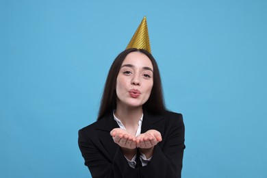 Happy woman in party hat blowing kiss on light blue background