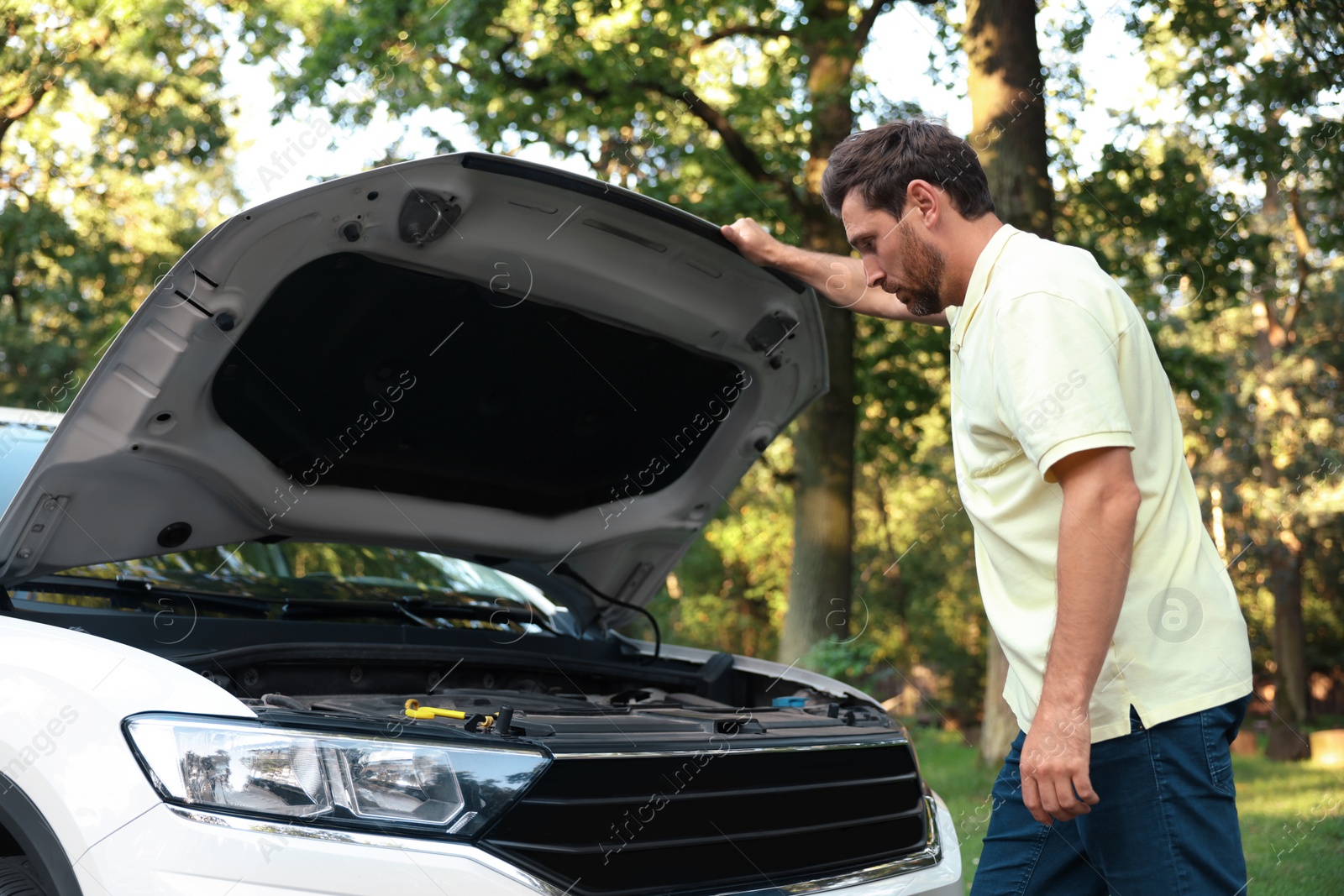 Photo of Upset man looking under hood of broken car outdoors