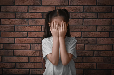 Photo of Sad little girl closing eyes with hands near brick wall. Child in danger