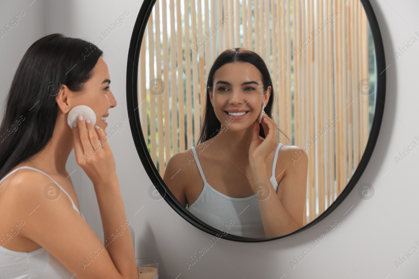 Photo of Young woman using cotton pad with micellar water near mirror indoors