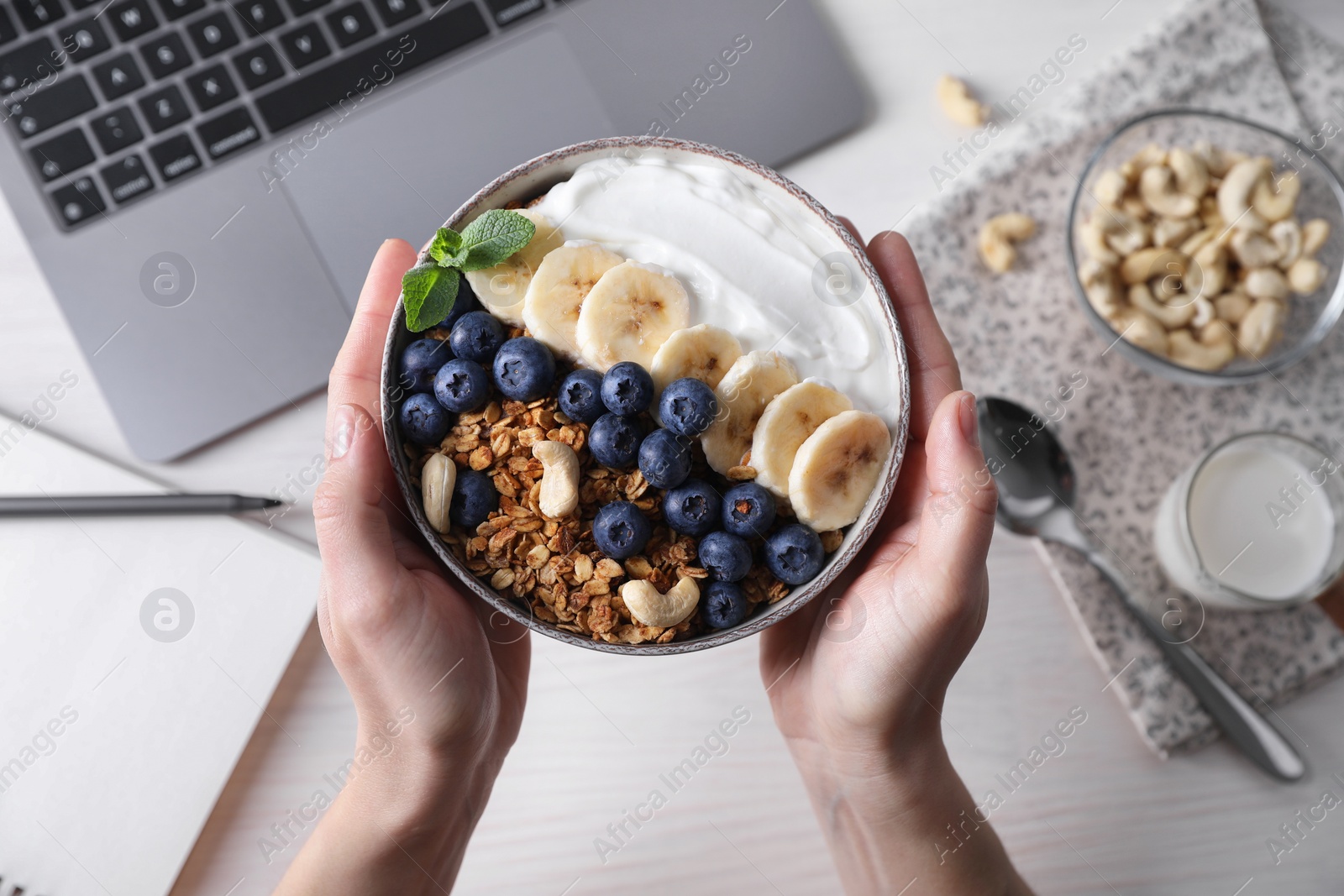 Photo of Woman holding bowl of tasty granola over white wooden table with laptop, top view