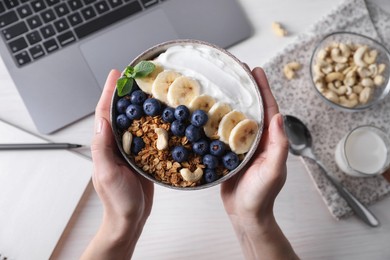 Woman holding bowl of tasty granola over white wooden table with laptop, top view