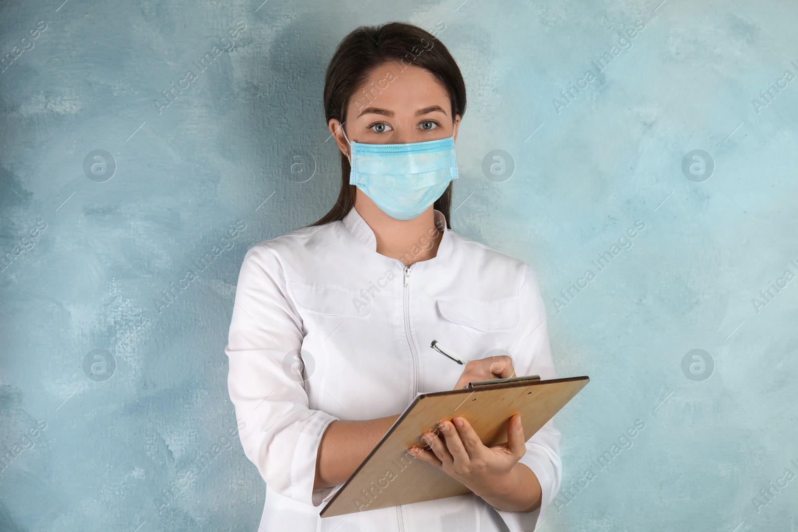 Photo of Doctor with disposable mask on face holding clipboard against light blue background