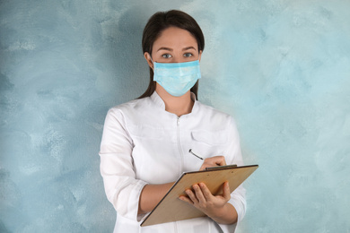 Photo of Doctor with disposable mask on face holding clipboard against light blue background