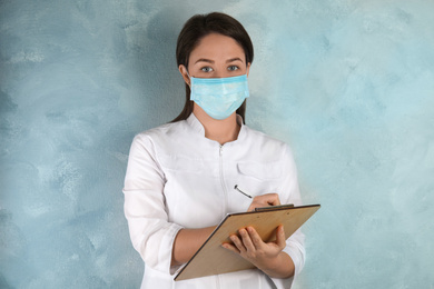 Doctor with disposable mask on face holding clipboard against light blue background