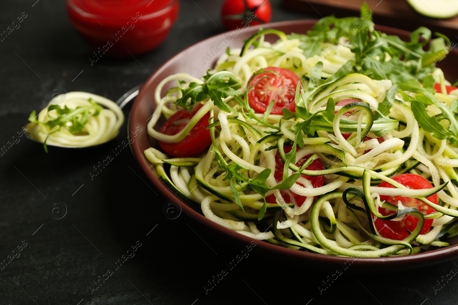 Photo of Delicious zucchini pasta with arugula and cherry tomatoes on black slate table, closeup