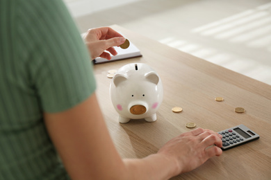 Photo of Woman putting money into piggy bank and using calculator at wooden table indoors, closeup