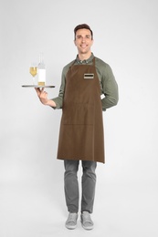 Handsome waiter holding tray with glass and bottle of wine on light background