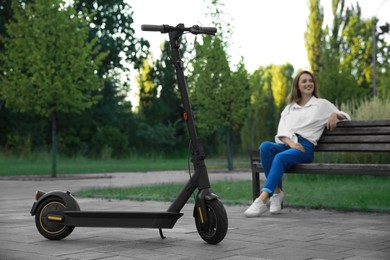 Photo of Modern electric kick scooter and woman sitting on bench in park, selective focus