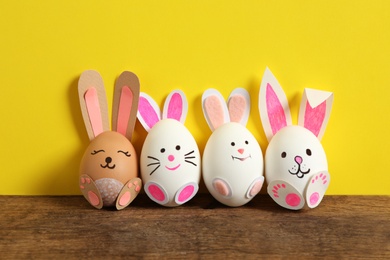 Eggs as cute bunnies on wooden table against yellow background. Easter celebration