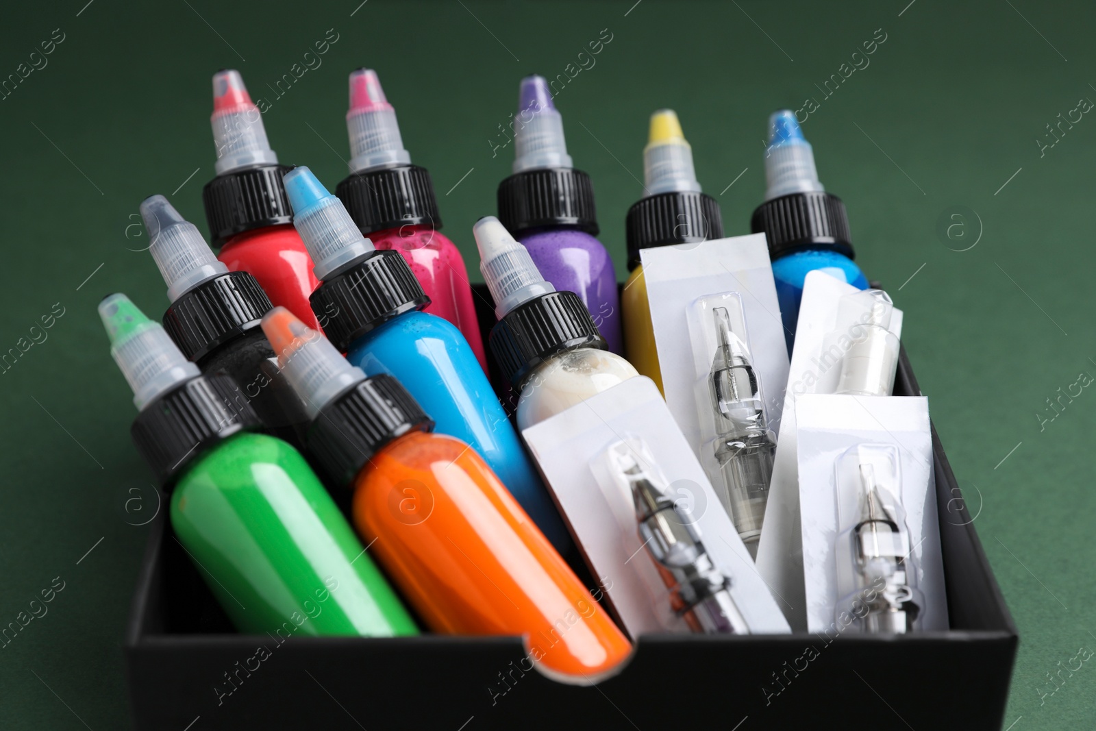 Photo of Box with colorful tattoo inks and needle cartridges on dark green background, closeup