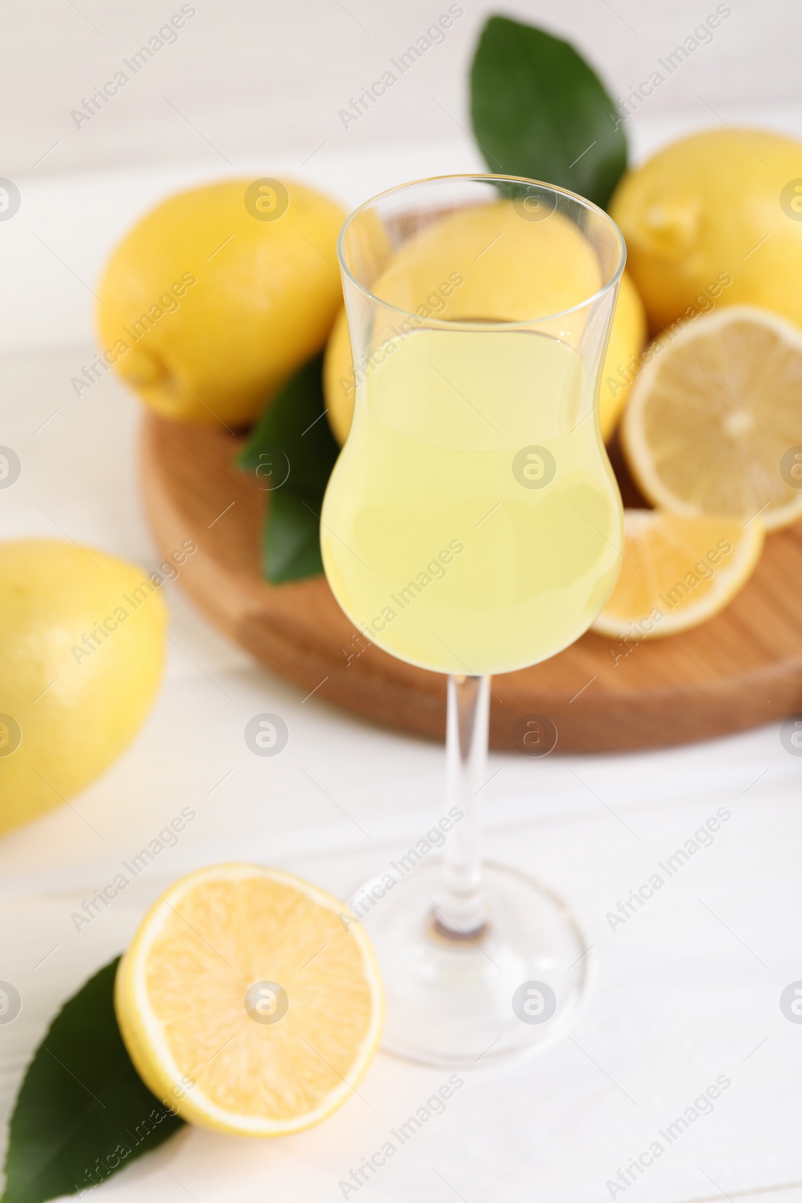 Photo of Liqueur glass with tasty limoncello, lemons and green leaves on white wooden table, closeup