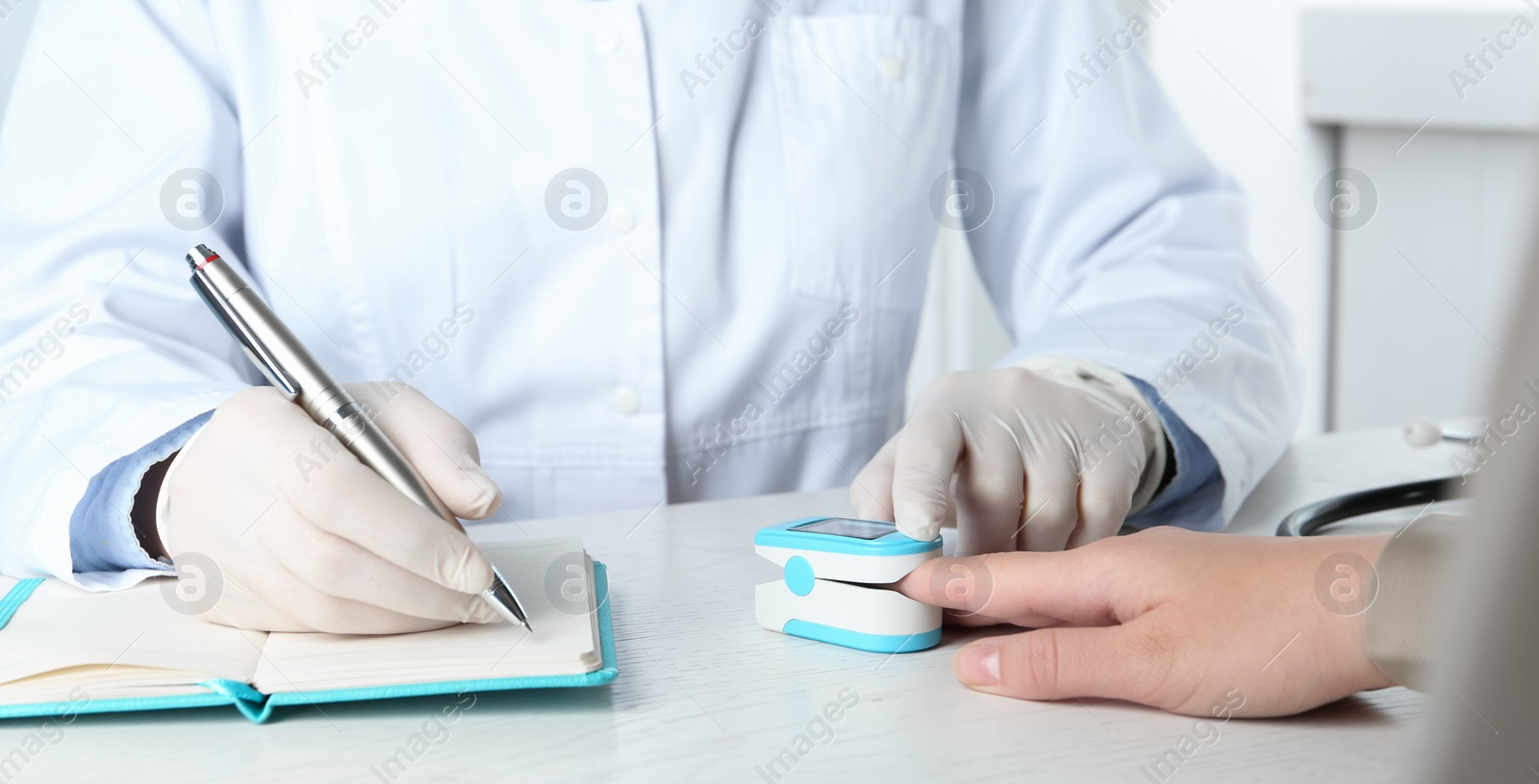 Photo of Doctor examining patient with fingertip pulse oximeter at white wooden table, closeup