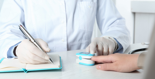 Photo of Doctor examining patient with fingertip pulse oximeter at white wooden table, closeup