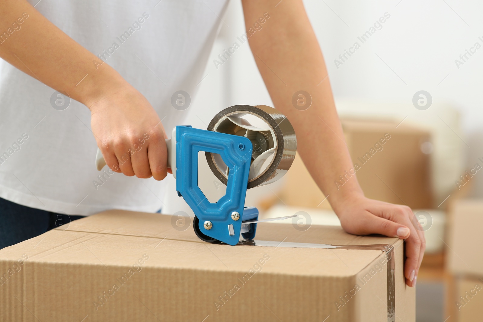 Photo of Woman applying adhesive tape on box with dispenser indoors, closeup