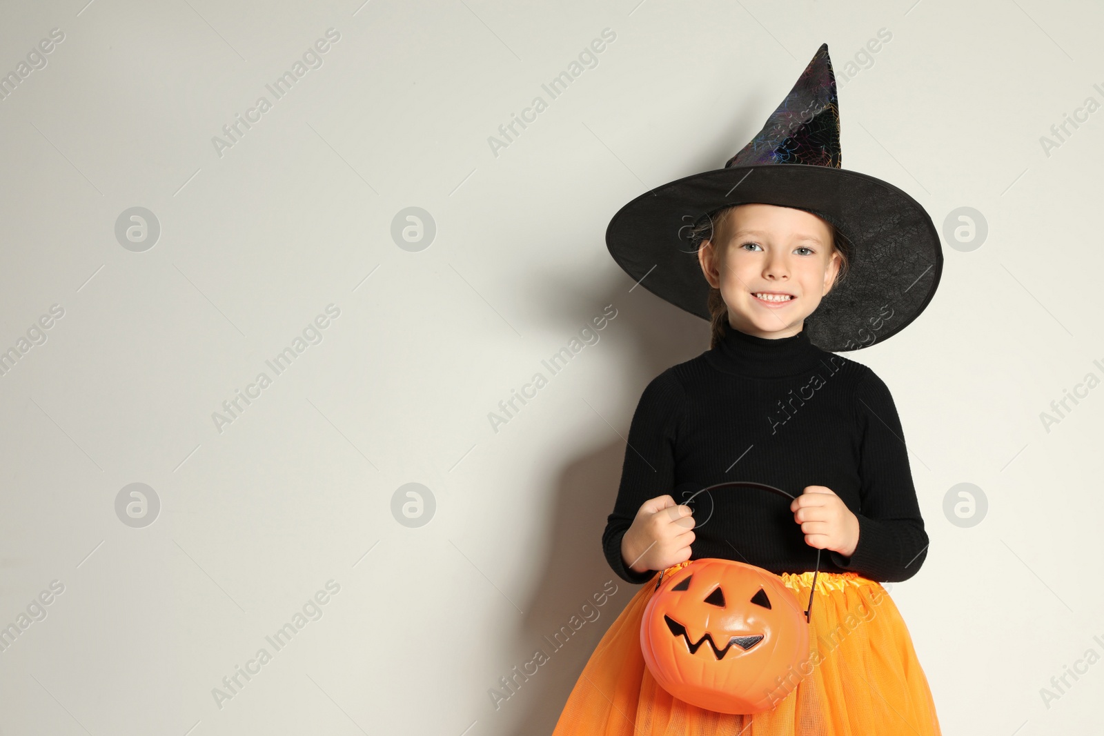 Photo of Cute little girl with pumpkin candy bucket wearing Halloween costume on light background. Space for text