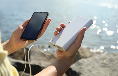 Photo of Woman charging smartphone with power bank on rocky mountain near river, closeup