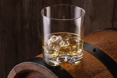 Whiskey with ice cubes in glass on barrel against wooden background, closeup