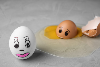 Photo of Whole and broken eggs with drawn faces on grey table. Concept of jealousy