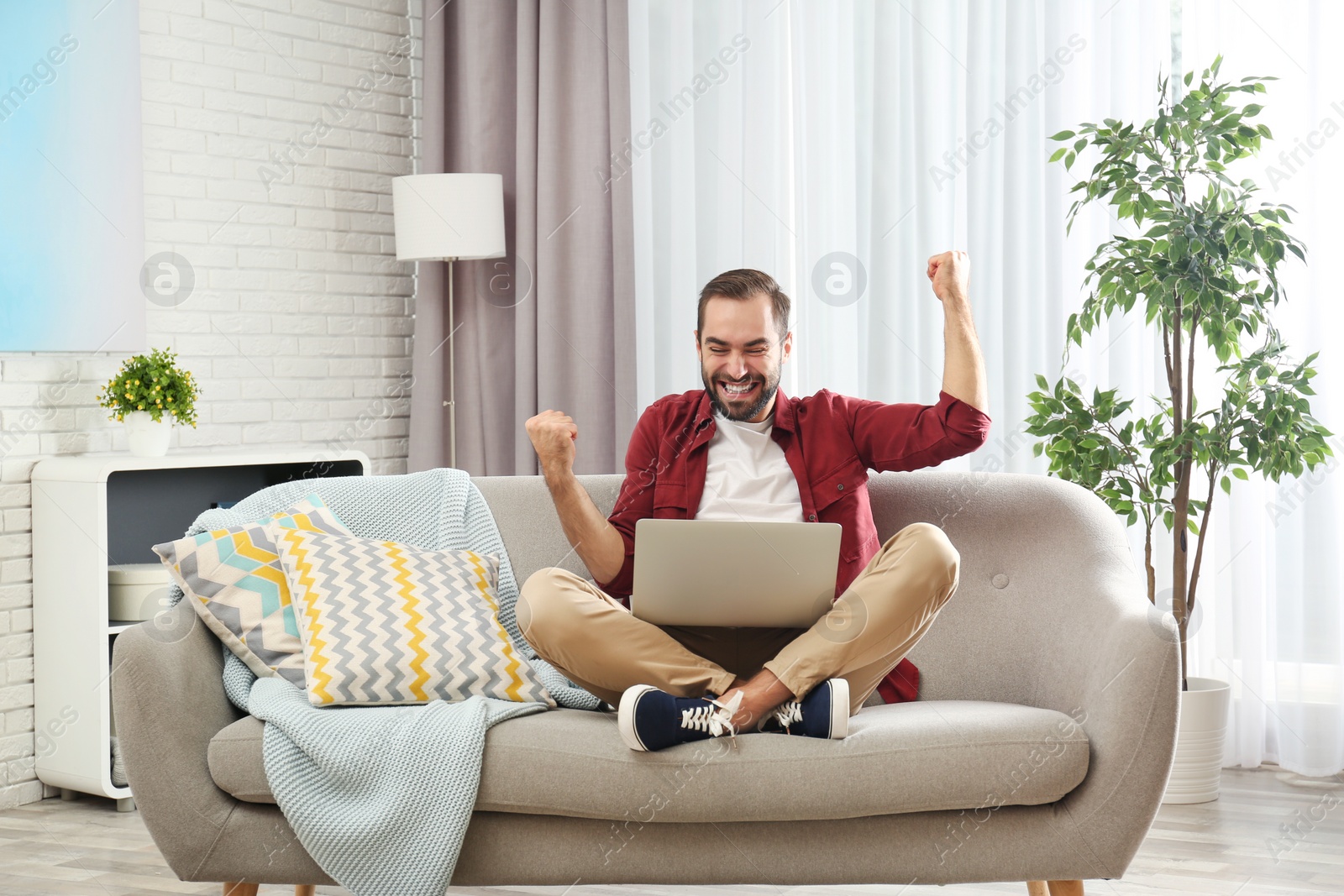 Photo of Emotional young man with laptop celebrating victory on sofa at home