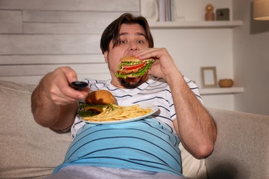 Photo of Overweight man with plate of burgers and French fries watching TV on sofa at home