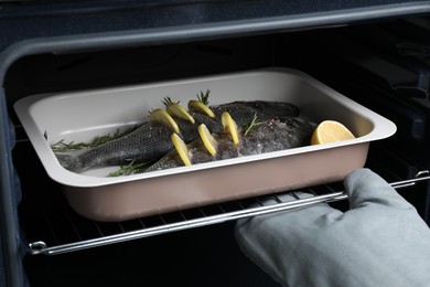 Woman putting baking tray with sea bass fish, lemon and rosemary into oven, closeup