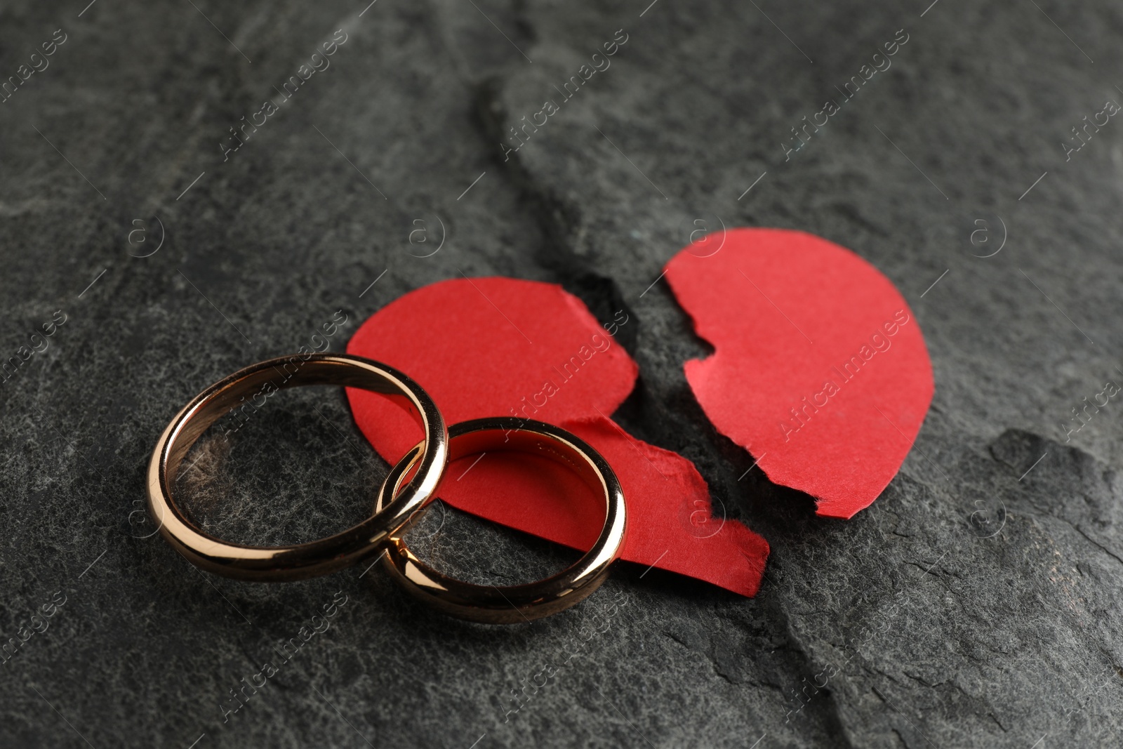 Photo of Halves of torn red paper heart and wedding rings on dark grey table. Broken heart