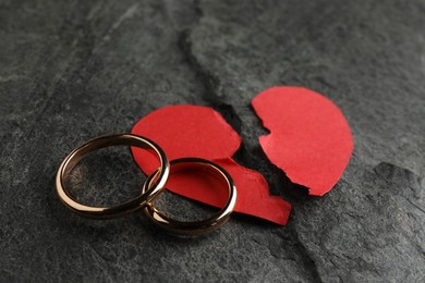 Photo of Halves of torn red paper heart and wedding rings on dark grey table. Broken heart