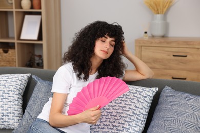 Young woman waving pink hand fan to cool herself on sofa at home