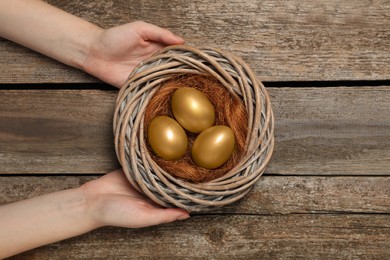 Woman holding nest with shiny golden eggs at wooden table, top view