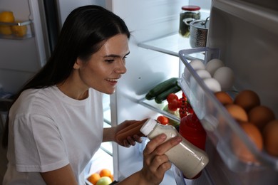 Young woman taking sauce and sausages out of refrigerator at night