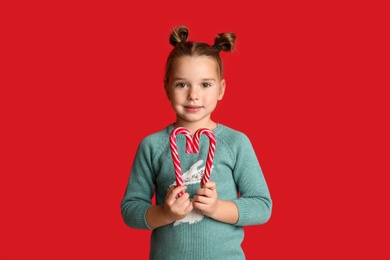 Photo of Cute little girl in knitted sweater holding candy canes on red background. Celebrating Christmas