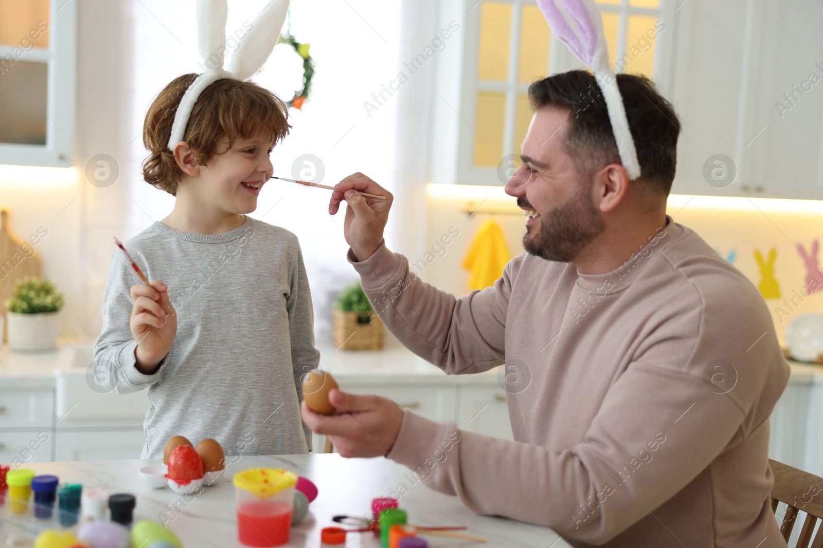 Photo of Easter celebration. Father with his little son having fun while painting eggs at white marble table in kitchen