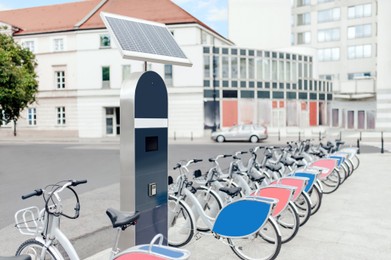 Photo of Station with solar panel and many bicycles on city street. Bike rental fleet