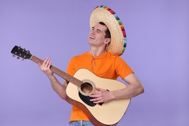 Young man in Mexican sombrero hat playing guitar on violet background