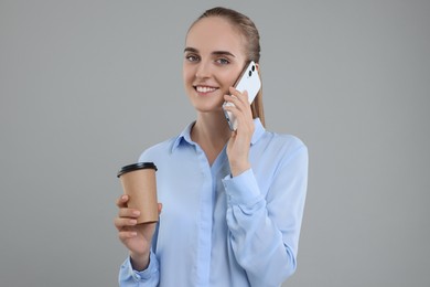 Photo of Happy young secretary with paper cup of drink talking on smartphone against grey background