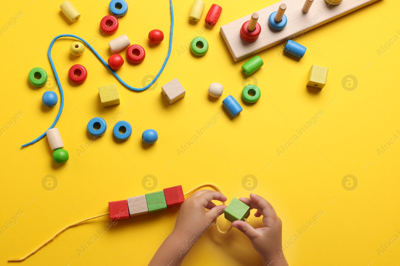 Photo of Motor skills development. Little child playing with wooden pieces and string for threading activity at yellow table, top view