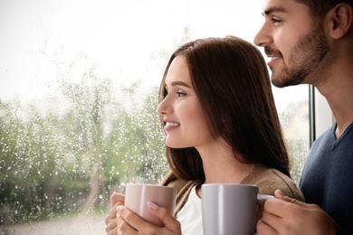 Photo of Happy young couple with cups near window indoors on rainy day