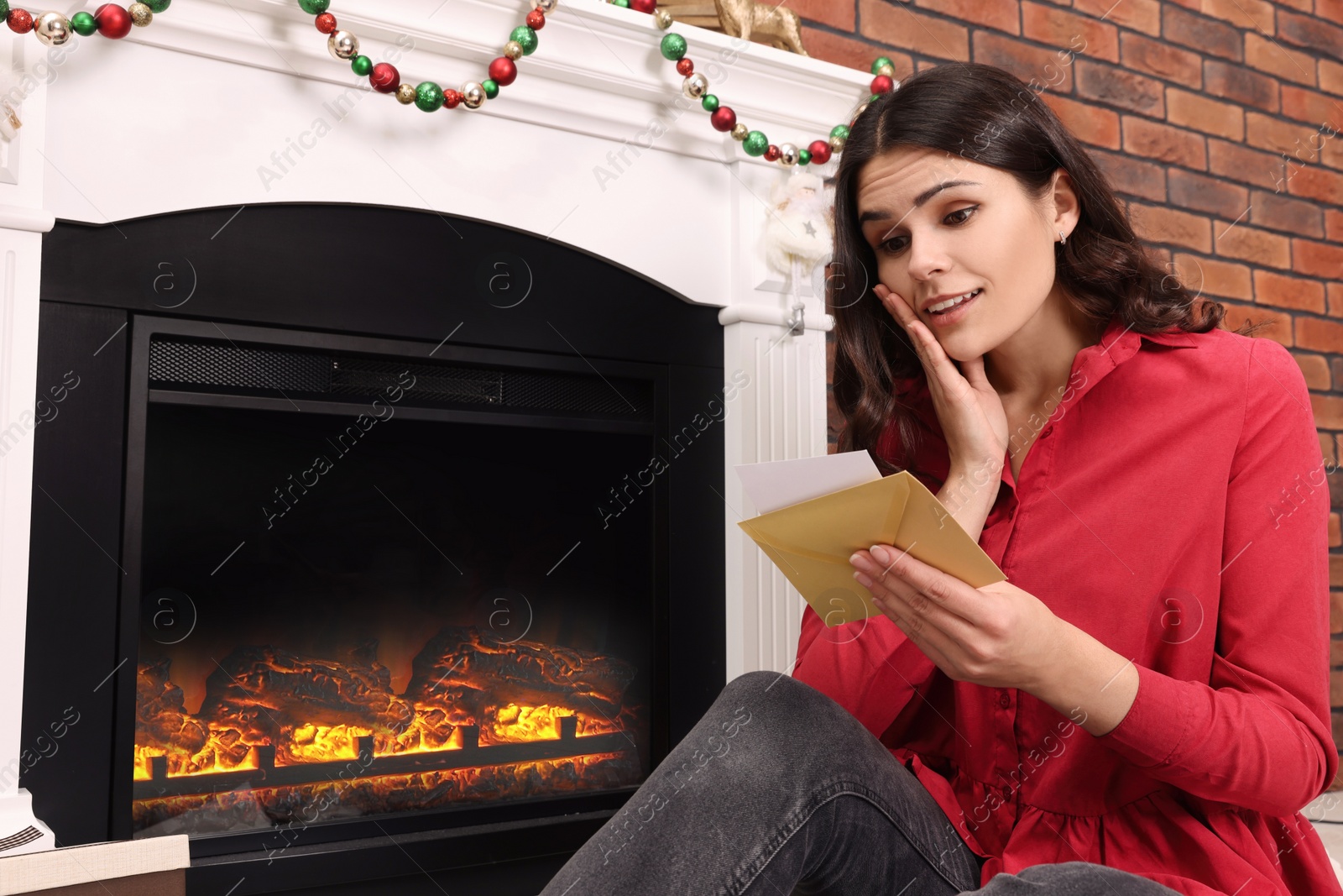 Photo of Emotional woman sitting near fireplace with greeting card indoors